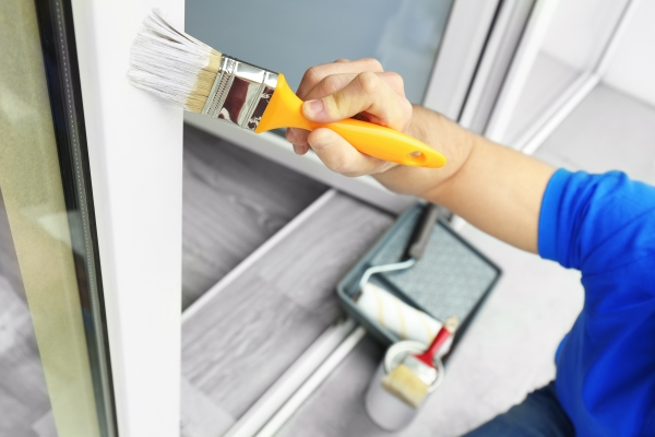 Closeup of a man using a paint brush to paint a window frame with white paint.
