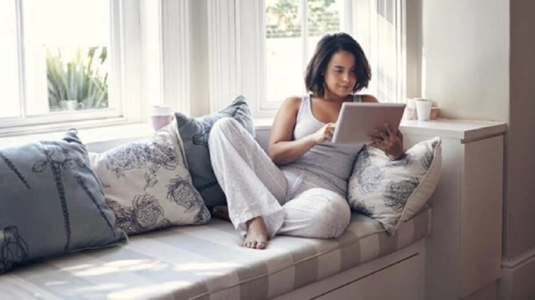 Woman looking at her tablet while lying on a window seat