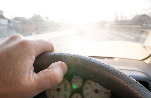 Person driving with a hand on the wheel and looking into the glare from the sun coming through a windshield