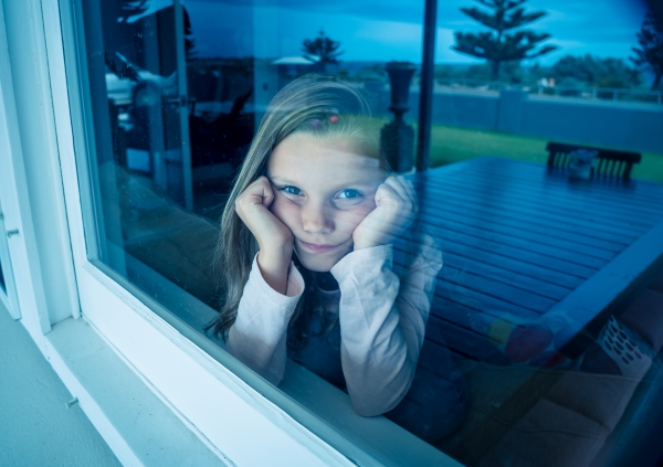 Young girl looking out window of her home while resting her chin on her hands.