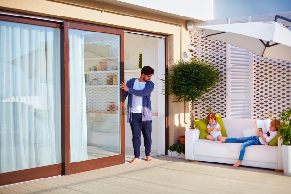 Young family members relaxing on outdoor patio, with man standing by sliding glass patio door.